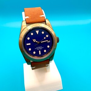 Pu&ograve; includere: Un orologio da polso color bronzo con quadrante blu e cinturino in pelle marrone. L'orologio ha indici e lancette bianche, con un'indicazione della data alle 3. &Egrave; esposto su un supporto bianco su sfondo blu.