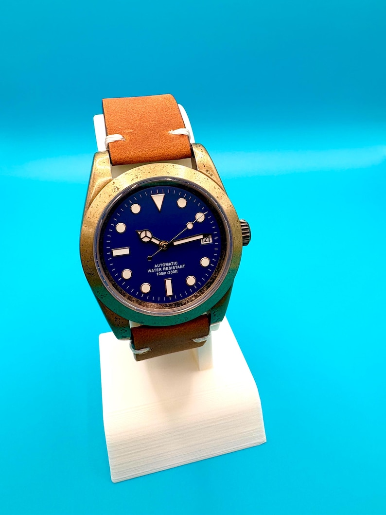 Pu&ograve; includere: Un orologio da polso color bronzo con quadrante blu e cinturino in pelle marrone. L'orologio ha indici e lancette bianche, con un'indicazione della data alle 3. &Egrave; esposto su un supporto bianco su sfondo blu.