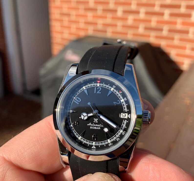 May include: A black and silver wristwatch with a black rubber band. The watch face has white markings and a small white square with the text 'Automatic 20BAR'.