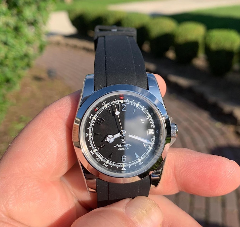 May include: A stainless steel wristwatch with a black dial and a black rubber strap. The watch face features white numerals, hands, and a date display. The watch is held in a hand against a blurred green background.