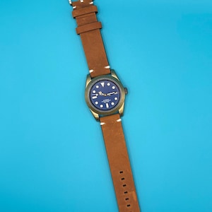Pu&ograve; includere: Un orologio da polso con cinturino in pelle marrone e quadrante blu. L'orologio ha una cassa color oro e cuciture bianche sul cinturino. L'orologio &egrave; esposto su sfondo blu.