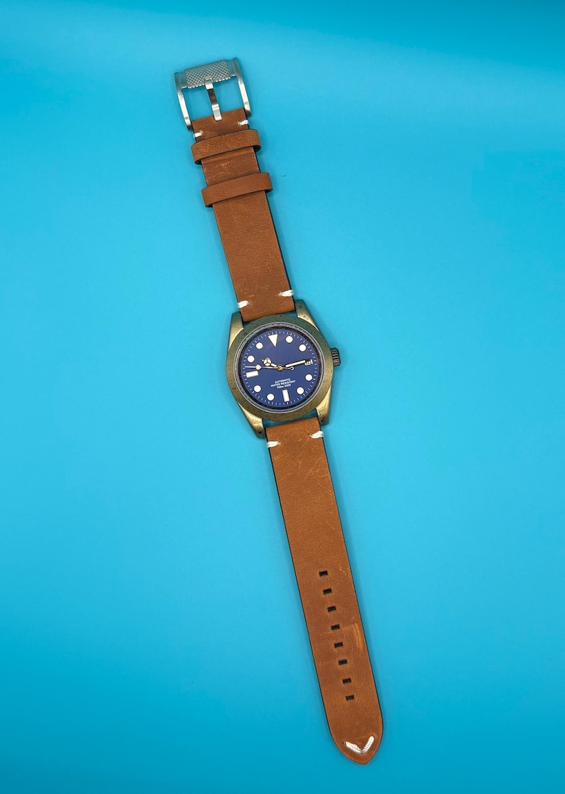 Pu&ograve; includere: Un orologio da polso con cinturino in pelle marrone e quadrante blu. L'orologio ha una cassa color oro e cuciture bianche sul cinturino. L'orologio &egrave; esposto su sfondo blu.
