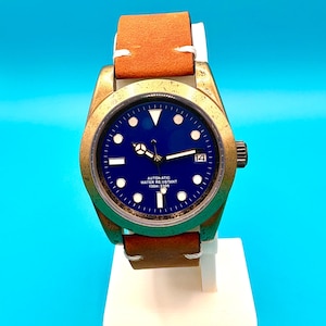 Pu&ograve; includere: Un orologio da polso con quadrante blu e cinturino in pelle marrone. L'orologio ha una cassa color oro e un'indicazione della data alle 3. L'orologio &egrave; contrassegnato "AUTOMATIC WATER RESISTANT 100m: 330ft".