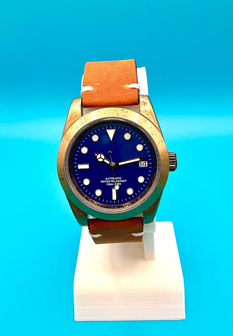 Pu&ograve; includere: Un orologio da polso con quadrante blu e cinturino in pelle marrone. L'orologio ha una cassa color oro e un'indicazione della data alle 3. L'orologio &egrave; contrassegnato "AUTOMATIC WATER RESISTANT 100m: 330ft".