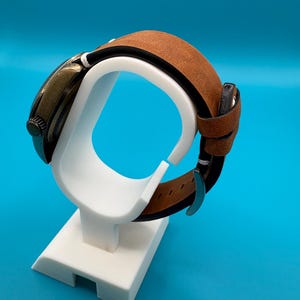 Pu&ograve; includere: Un orologio in stile vintage con cinturino in pelle marrone &egrave; esposto su un supporto bianco. L'orologio ha un quadrante scuro e un cinturino in pelle marrone. Il supporto &egrave; bianco e ha un design semplice. Lo sfondo &egrave; blu.