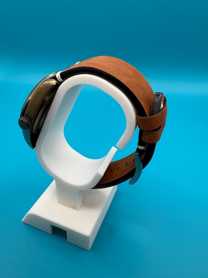 Pu&ograve; includere: Un orologio in stile vintage con cinturino in pelle marrone &egrave; esposto su un supporto bianco. L'orologio ha un quadrante scuro e un cinturino in pelle marrone. Il supporto &egrave; bianco e ha un design semplice. Lo sfondo &egrave; blu.