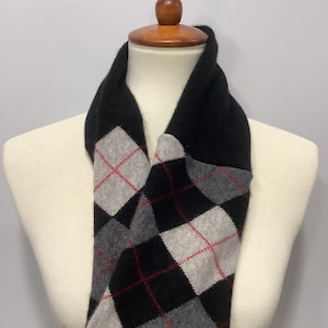 May include: A black and gray argyle infinity scarf with red accents is displayed on a mannequin. The scarf is made of a soft material and is designed to be worn around the neck. The argyle pattern is a classic design.