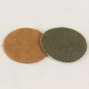 May include: Two small, round felt pads. One is a light brown color, and the other is a muted olive green. The pads are positioned close together against a white background. These could be used for crafts or as furniture protectors.