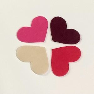 May include: Four heart-shaped felt cutouts in shades of pink, burgundy, red, and cream, arranged in a cloverleaf pattern against a white background. The felt has a soft, textured appearance.