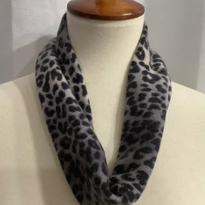 May include: A soft infinity scarf featuring a grey and black leopard print pattern. The fabric displays a continuous loop design with a fuzzy texture, showcasing dark charcoal spots against a lighter grey background throughout the entire garment.