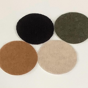 May include: Four circular felt coasters arranged in a cluster. The set includes one solid black coaster, one dark olive green coaster, one medium brown coaster, and one light beige coaster. Each piece features a flat, uniform, and textured surface.