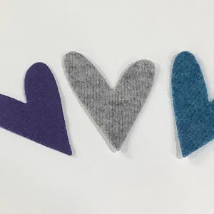 May include: Three heart-shaped fabric cutouts arranged in a row. The left heart is deep purple, the center heart is heather gray, and the right heart is a textured teal blue. Each heart features a distinct, slightly asymmetrical shape.