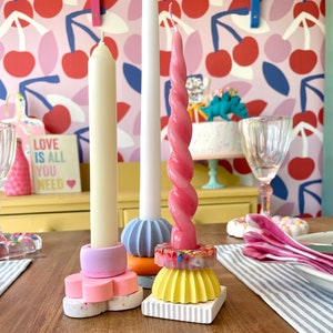 Puede incluir: Un conjunto de velas y candelabros coloridos sobre una mesa de madera. Las velas son crema, blancas y rosas, con un diseño retorcido. Los candelabros son de varias formas y colores, incluyendo rosa, azul, naranja y amarillo. Un letrero dice "LOVE IS ALL YOU NEED."