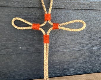 Könnte beinhalten: Ein handgefertigtes Kreuz aus Naturseil mit orangefarbenen Bindfäden an den Verbindungsstellen und Enden. Das Kreuz wird vor einem dunkelblauen Holzgrund präsentiert, der seine rustikale Textur und sein Design hervorhebt.
