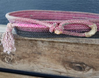 Könnte beinhalten: Ein ovaler Korb aus gewickeltem Seil in den Farben Pink, Magenta und Schwarz. Der Rand ist mit einem dekorativen geknoteten Seildetail versehen, an dessen einer Seite eine ausgefranste Quaste hängt und an der anderen ein heller Akzent.