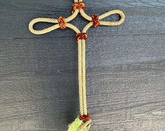Könnte beinhalten: Ein handgefertigtes Kreuz aus beigem Seil mit aufwendigen Knoten und Akzenten aus braunem Seil. Das Kreuz hat oben ein Schlaufendesign und unten eine Quaste aus grünen und beigen Fasern. Der Hintergrund ist eine dunkelblaue Holzoberfläche.