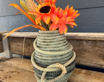 Könnte beinhalten: Eine dekorative Vase aus gewickeltem Seil, die ein lebendiges Arrangement aus orangefarbenen Sonnenblumen und Weizenähren enthält. Die Vase hat eine einzigartige Textur und ein geknotetes Seildetail, das eine rustikale, nautische Ästhetik schafft.