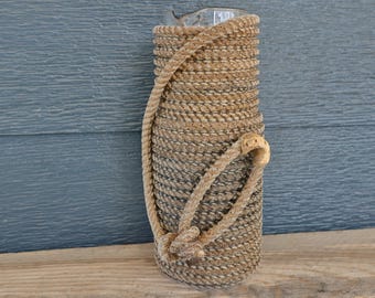 Könnte beinhalten: Eine zylindrische Vase aus klarem Glas, die mit dickem, beigefarbenem Naturfaserseil umwickelt ist. Das Seil ist fest um die gesamte Außenseite des Glases gewickelt, und ein separates Seilstück ist zu einer dekorativen Schlaufe an der Seite geknotet.