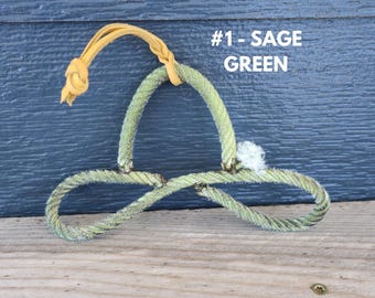 Könnte beinhalten: Ein salbeigrünes Seilornament in Form einer stilisierten Schleife oder eines Knotens mit einer hellbraunen Lederschnur zum Aufhängen. Der Text #1 - SAGE GREEN ist in Weiß über dem Seilobjekt mit seiner strukturierten, faserigen Oberfläche gedruckt.