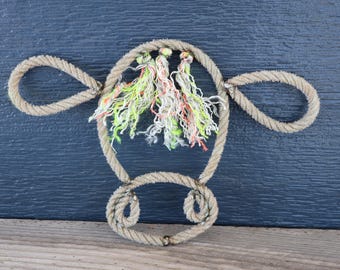 Könnte beinhalten: Ein Wandbehang aus beigefarbenem, gedrehtem Seil in Form eines Kuhkopfes mit zwei seitlichen Schlaufen als Ohren und einer unteren Schlaufe als Schnauze. Drei Quasten aus neongelbem, orangefarbenem und weißem Fransen-Garn hängen oben mittig.