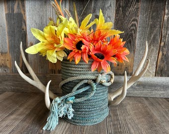 Könnte beinhalten: Eine dekorative Vase, umwickelt mit blauem Seil, gefüllt mit gelben und orangefarbenen Kunstblumen. Die Vase wird von zwei weißen Hirschgeweihen flankiert, wodurch ein rustikales, ländliches Blumenarrangement entsteht.