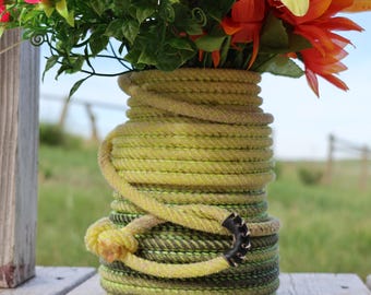 Könnte beinhalten: Eine zylindrische Vase, die mit dickem, gewickeltem Seil ummantelt ist und einen Farbverlauf von Limettengrün bis Dunkelolivgrün aufweist. Ein loses Seilstück mit einem schwarz vernähten Lederende und einem Knoten ist um die Vase drapiert.