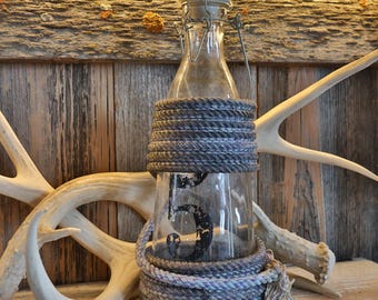Könnte beinhalten: Eine klare Glasflasche mit Bügelverschluss, die auf der Vorderseite mit einer schwarzen Ziffer 5 bedruckt ist. Die Flasche ist im oberen und unteren Bereich mit einem dicken, strukturierten grauen Seil umwickelt.