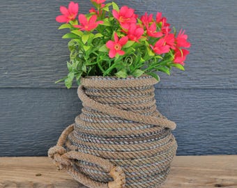 Könnte beinhalten: Eine Vase aus gewickeltem braunem Seil mit strukturierter Oberfläche, die an der Seite einen dekorativen Seilgriff aufweist. Die Vase ist mit einem Strauß leuchtend rosa Blumen mit gelben Mitten und grünen Blättern gefüllt, die einen Kontrast bilden.