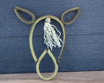 Könnte beinhalten: Ein Wandbehang in Form eines Kuhkopfes, gefertigt aus dickem, hellem gedrehtem Seil. Das Design besteht aus zwei spitzen Ohren, einem abgerundeten Gesicht und einem kleineren Nasenbereich, akzentuiert durch eine weiße, ausgefranste Quaste.
