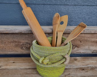 Könnte beinhalten: Ein aus limegrünem Seil gewickelter Korb mit einem hölzernen Nudelholz und drei hölzernen Küchenutensilien. Der Korb verfügt über ein geknotetes Seildetail auf der Vorderseite und ein kleines halbmondförmiges Akzentstück an der Seite der Seilwicklung.