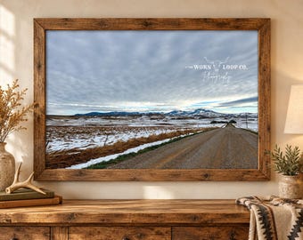 Könnte beinhalten: Ein gerahmtes Landschaftsfoto mit einem Feldweg, der unter einem bewölkten Himmel auf schneebedeckte Berge zuführt. Das Bild enthält das weiße Textlogo THE WORN LOOP CO. Photography mit einem Stierschädel-Symbol oben rechts.