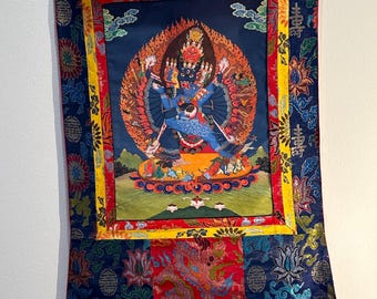 May include: A colorful Tibetan Buddhist thangka depicting a deity with multiple arms and heads, surrounded by flames and other figures. The thangka is framed with a yellow, red, and blue border and has a floral pattern on the bottom.
