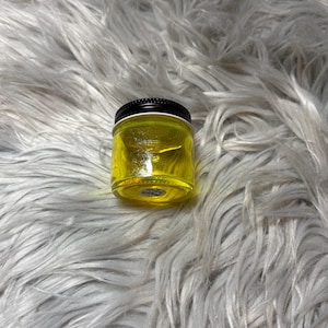 May include: A small, clear glass jar filled with a bright yellow liquid. The jar has a black lid and a small, round label on the bottom. The jar is sitting on a fluffy, white surface.
