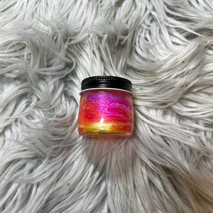May include: A small, square glass jar with a black lid. The jar's contents display a vibrant blend of colors, including pink, orange, and gold, creating a sunset-like effect. The jar is set against a fluffy, gray background.