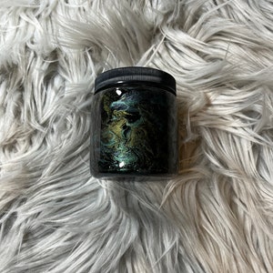 May include: A black glass jar with a black lid filled with a dark green and gold glitter mixture.