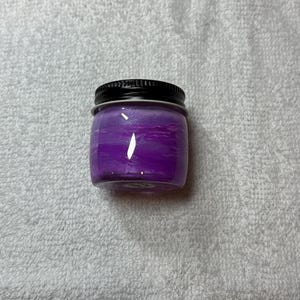 May include: A small, clear glass jar with a black lid. The jar is filled with a vibrant purple substance, with a marbled effect. The jar is sitting on a white, textured surface.
