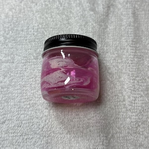 May include: A small, clear glass jar with a black lid, filled with a pink and white swirled substance. The jar sits on a white, textured surface. The jar appears to be a container for a cosmetic or craft product.