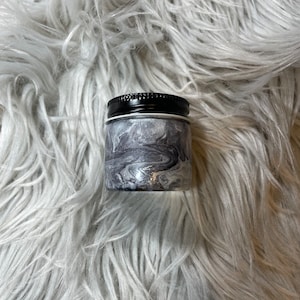 May include: A small, round, black and white marbled jar with a black lid. The jar is sitting on a white fluffy surface.