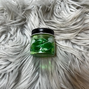 May include: A small glass jar with a black lid filled with green glitter. The jar is sitting on a white fluffy surface.