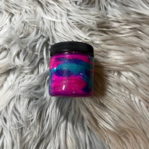 May include: A small glass jar with a black lid filled with pink, blue, and purple glitter. The jar is sitting on a fluffy white surface.