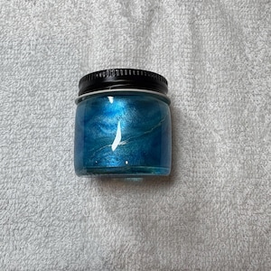May include: A small, clear glass jar with a black lid. The jar is filled with a shimmering, blue substance. The jar is sitting on a white, textured surface.