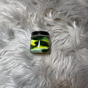 May include: A small, square jar with a black lid. The jar's contents feature a vibrant swirl of yellow and green colors. The jar is set against a backdrop of soft, gray, faux fur.