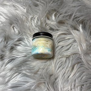 May include: A small, clear glass jar with a black lid. The jar contains a swirling mixture of light blue, gold, and white colors. The jar is sitting on a fluffy, white surface.