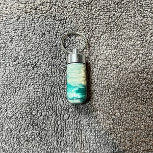 May include: A small, silver, cylindrical keychain with a teal, white, and green marbled design.
