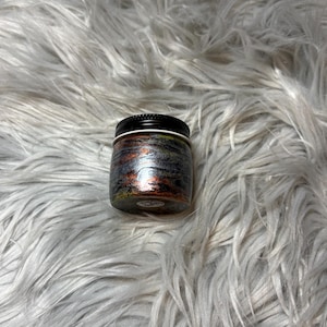 May include: A small, round jar with a black lid. The jar's exterior features a swirling pattern of metallic colors, including silver, copper, and black. The jar is sitting on a fluffy, white surface.