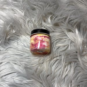 May include: A small, round jar with a black lid. The jar's contents display a swirl of pink, yellow, and white colors. The jar is set against a backdrop of fluffy, white faux fur.