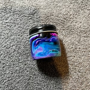 May include: A small, round jar with a black lid. The jar is decorated with a swirling pattern of blue, purple, and pink paint.