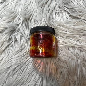 May include: A small glass jar with a black lid filled with a red and yellow liquid. The liquid has a swirling pattern.