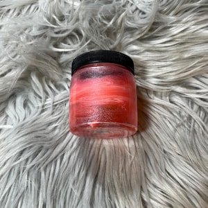 May include: A small, clear glass jar with a black lid. The jar is filled with a red, glittery substance that appears to be a cosmetic product. The contents have a layered effect, with a gradient of colors. The jar is sitting on a fluffy, gray surface.