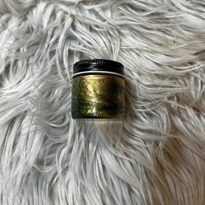 May include: A small, round jar with a black lid. The jar's exterior features a gold and green design. The jar is set against a backdrop of fluffy, gray faux fur.
